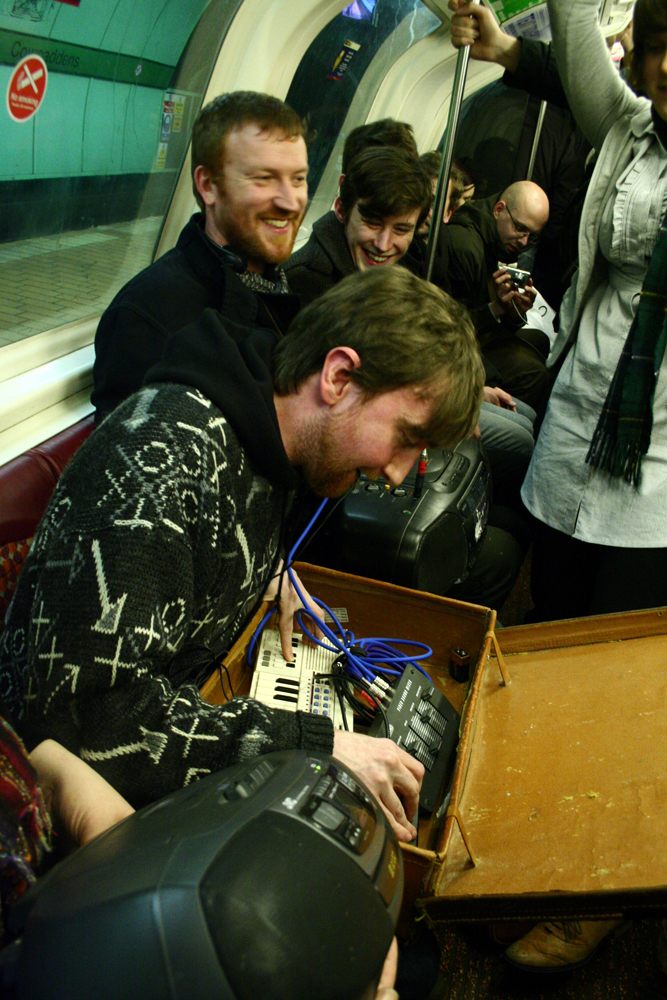 A performer plays instruments in a suitcase on a subway carriage