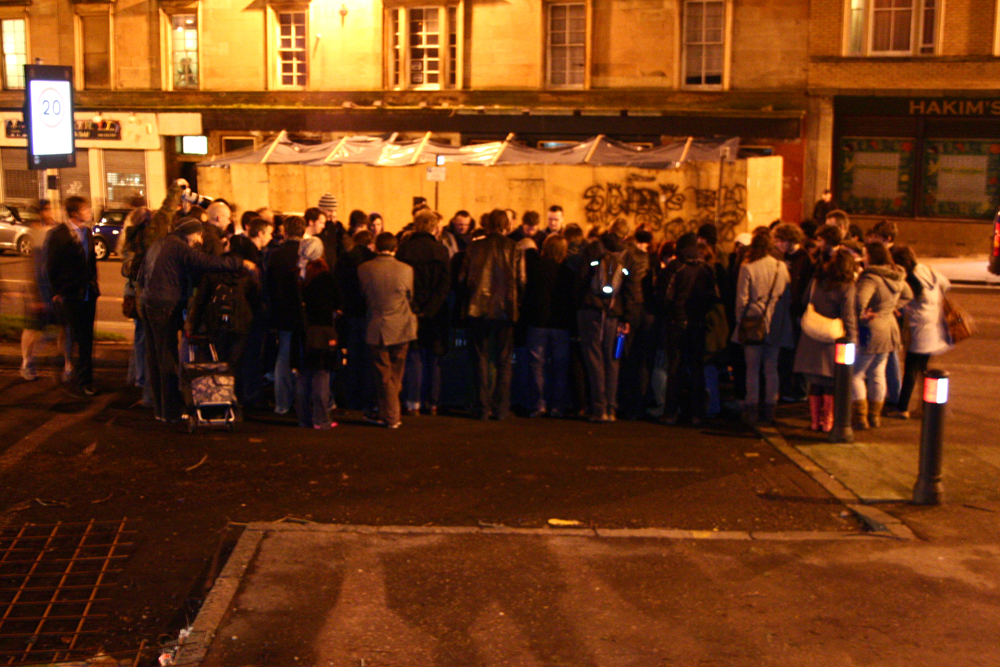 A crowd gathers around a skip by streetlight