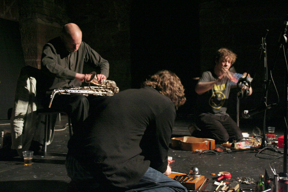 Three men play instruments in various stages of dismantlement