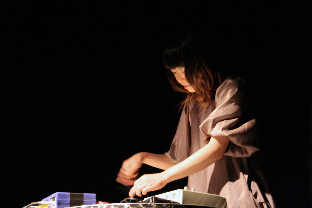 A performer leans over a mixer, they are lit but the space is dark