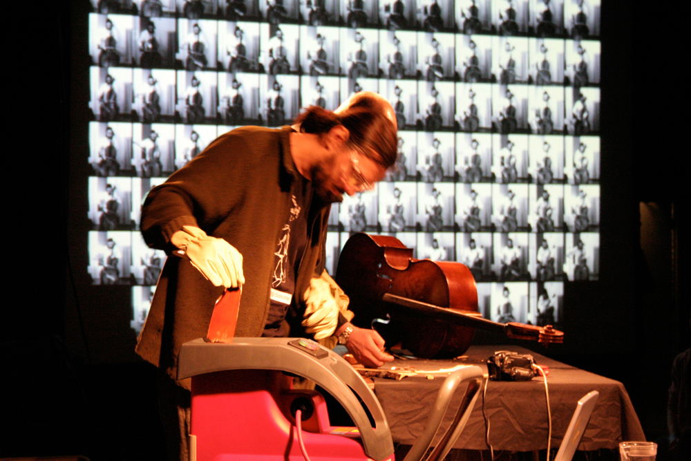 Nikos Veliotis in safety goggles chips wood, cello and a projection in the back