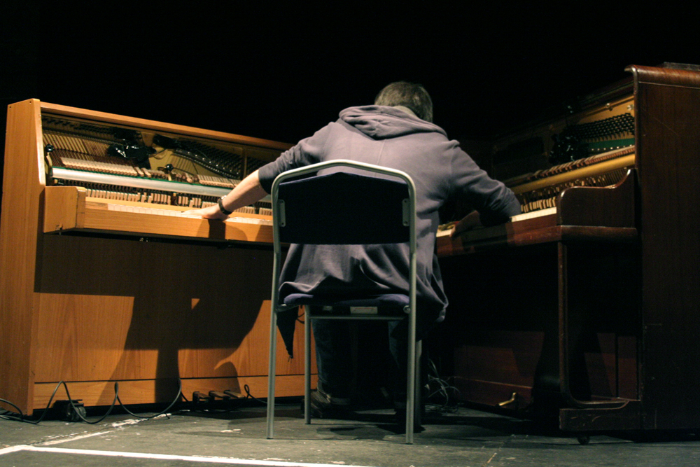 Otomo Yoshihide seated between two upright pianos their interiors exposed