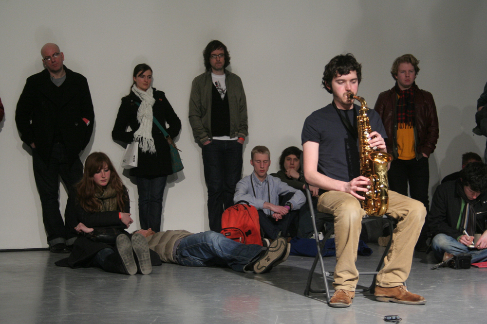 Marc Baron plays saxophone sat on a chair, an audience around