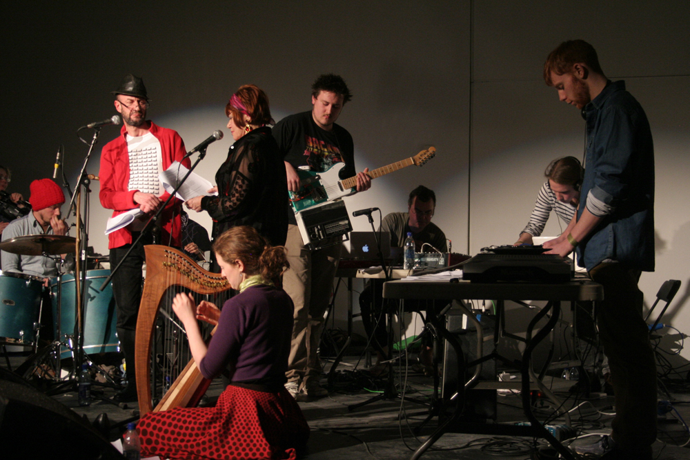 A band on stage including a harpist, a reader in a red shirt and electronics