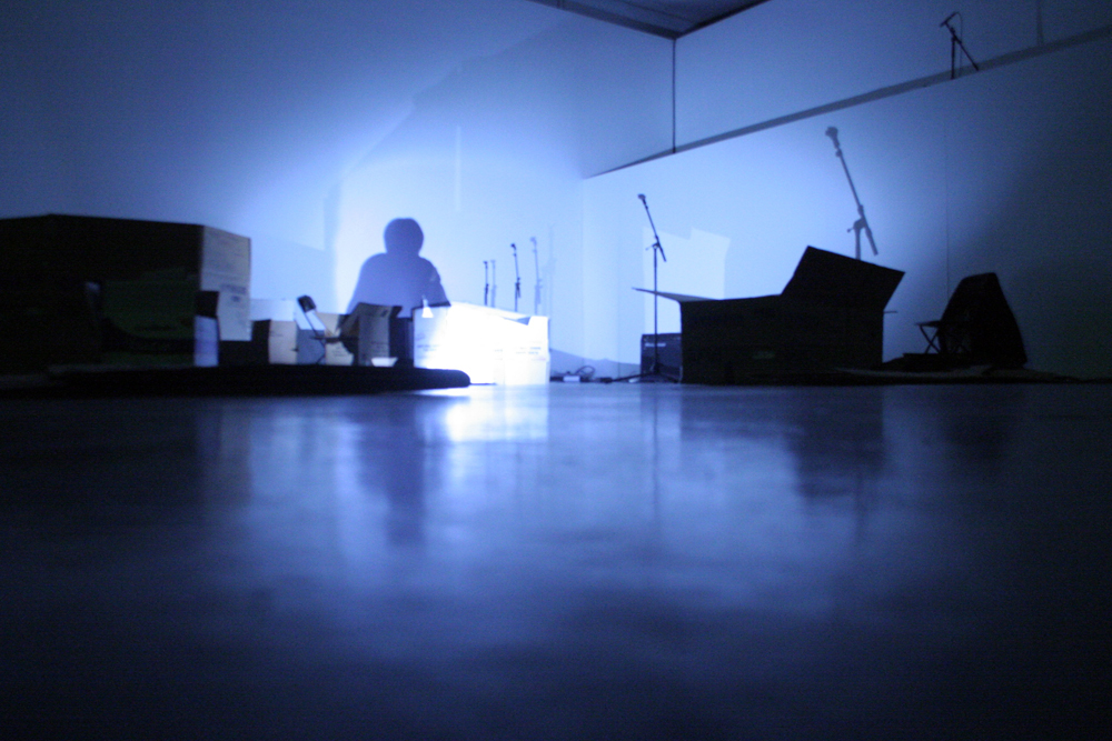A shadow crouches on the floor amongst mic stands and boxes, one light source