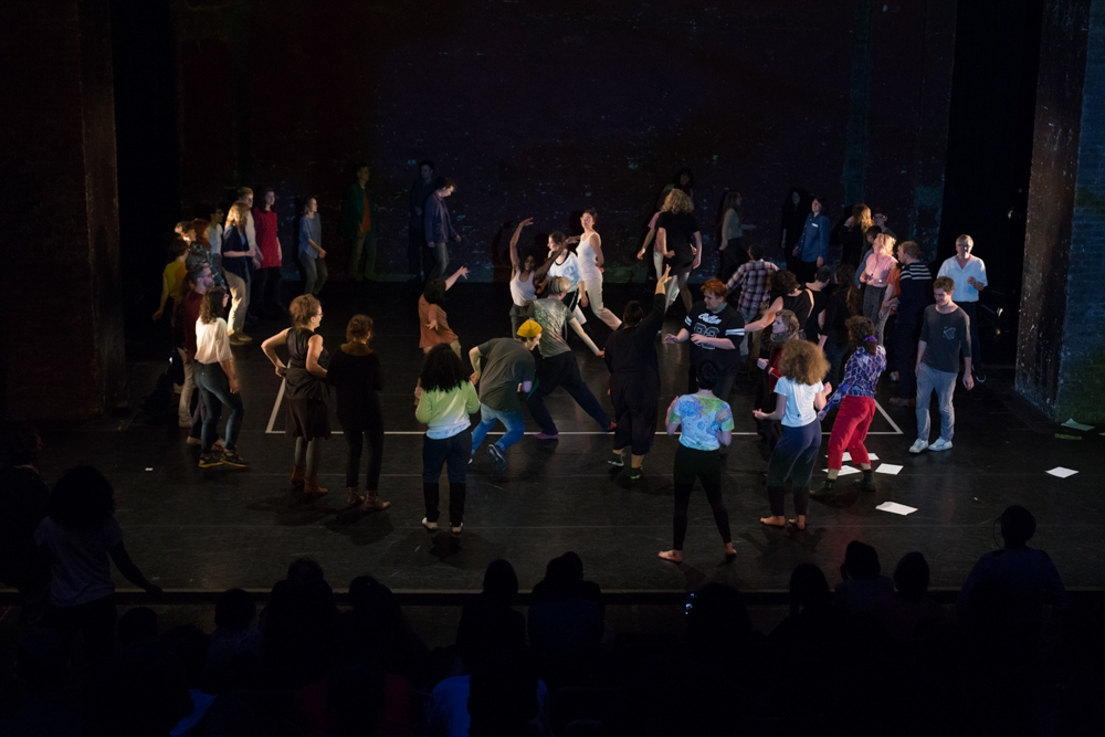 A large group of people on a stage dance and move aroud