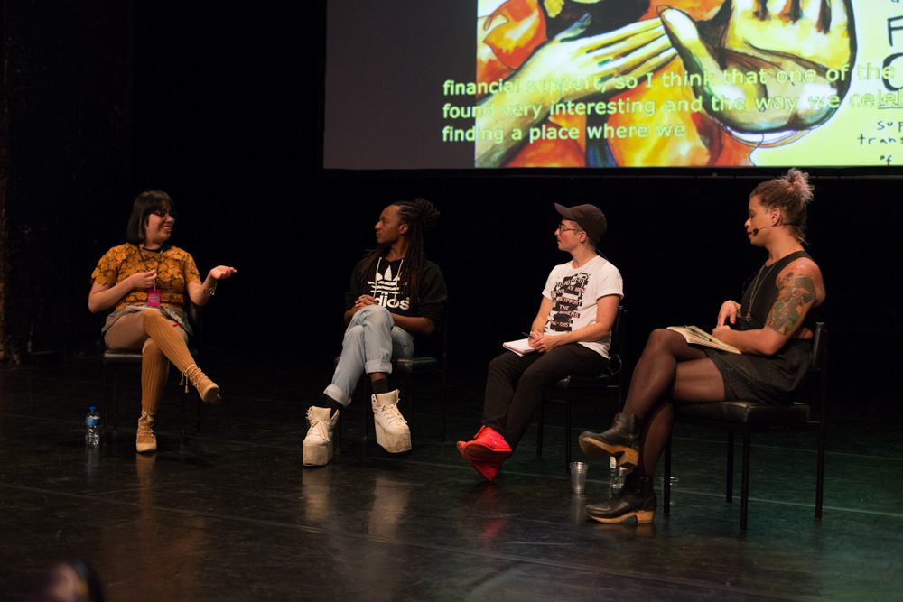 Four people on stage have a discussion. A screen projects images behind them