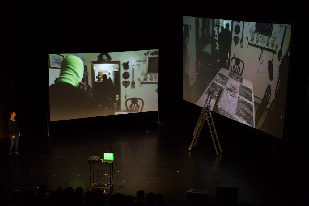 Two large screens on stage. Images of a dining table, someone wears a balaclava