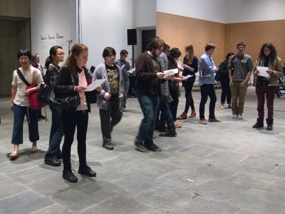 A group of people each holding a sheet of paper walk around each other