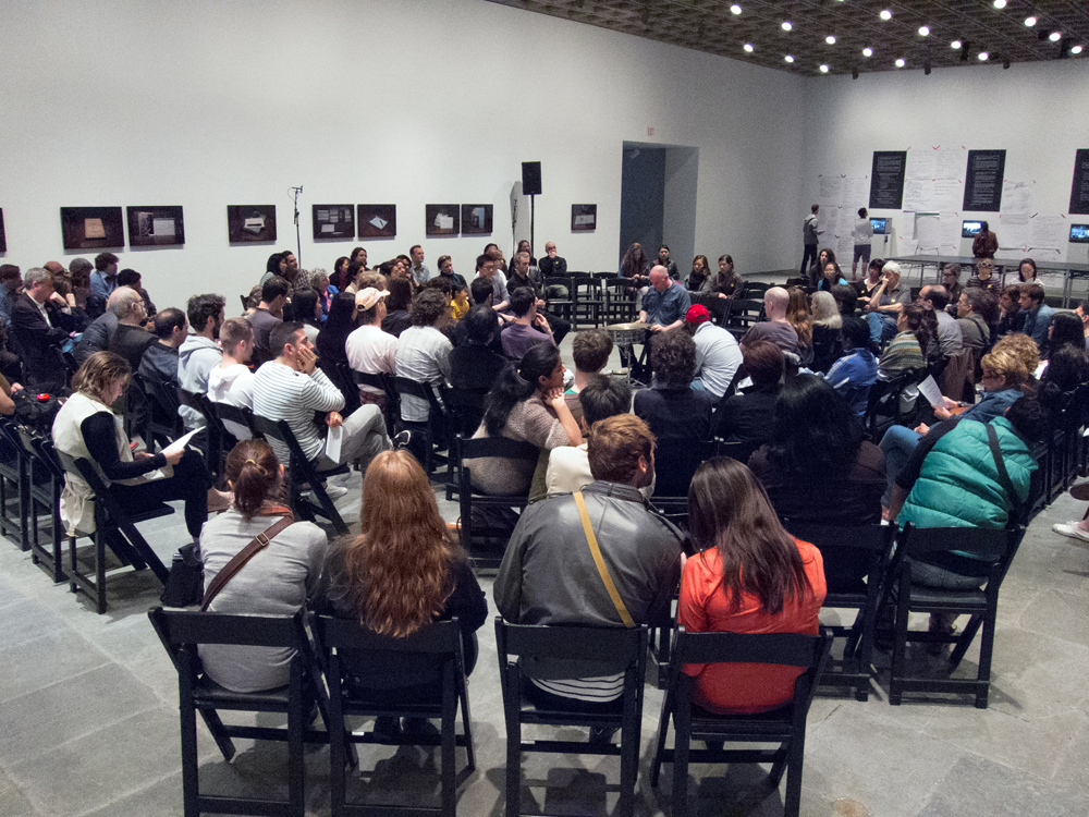A audience sat on black wooden chairs in a large gallery watches Sean Meehan