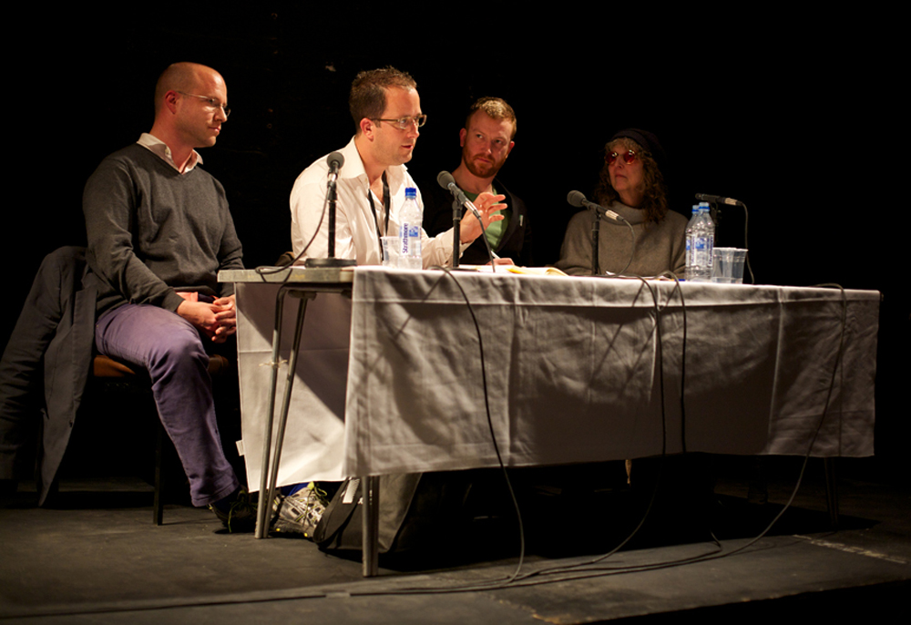 Robin MacKay sits on a panel of 4 people. He leans forward to speak into a mic