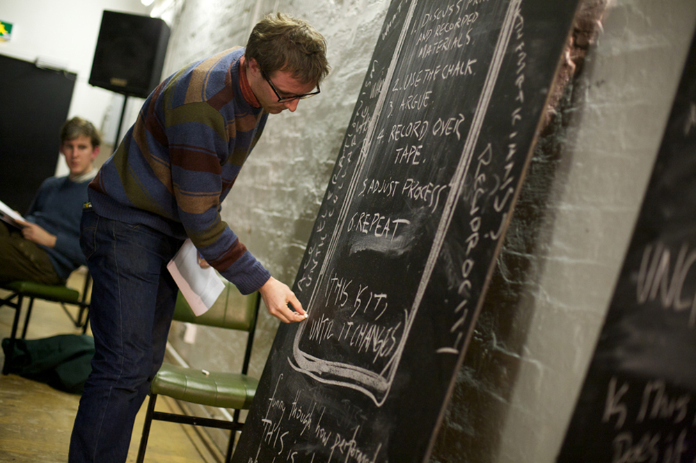 A man in a jumper writes on a large vertical blackboard