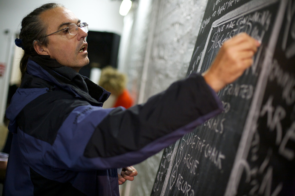 a man in glasses and a jacket stretches to write on a vertical blackboard