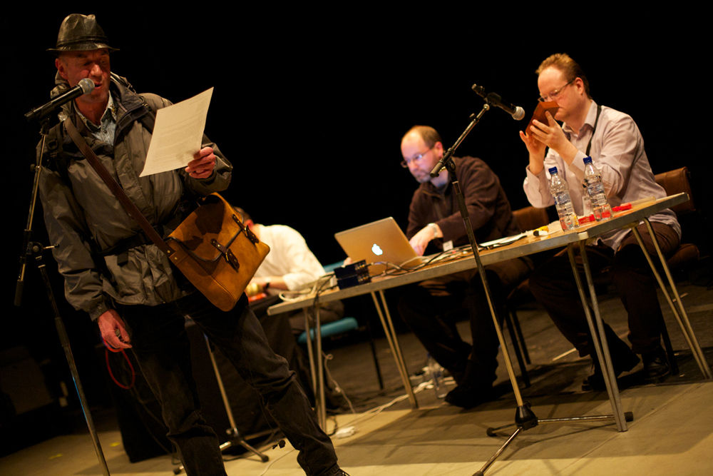 Tam Dean Burn in a hat reads from a paper into a microphone