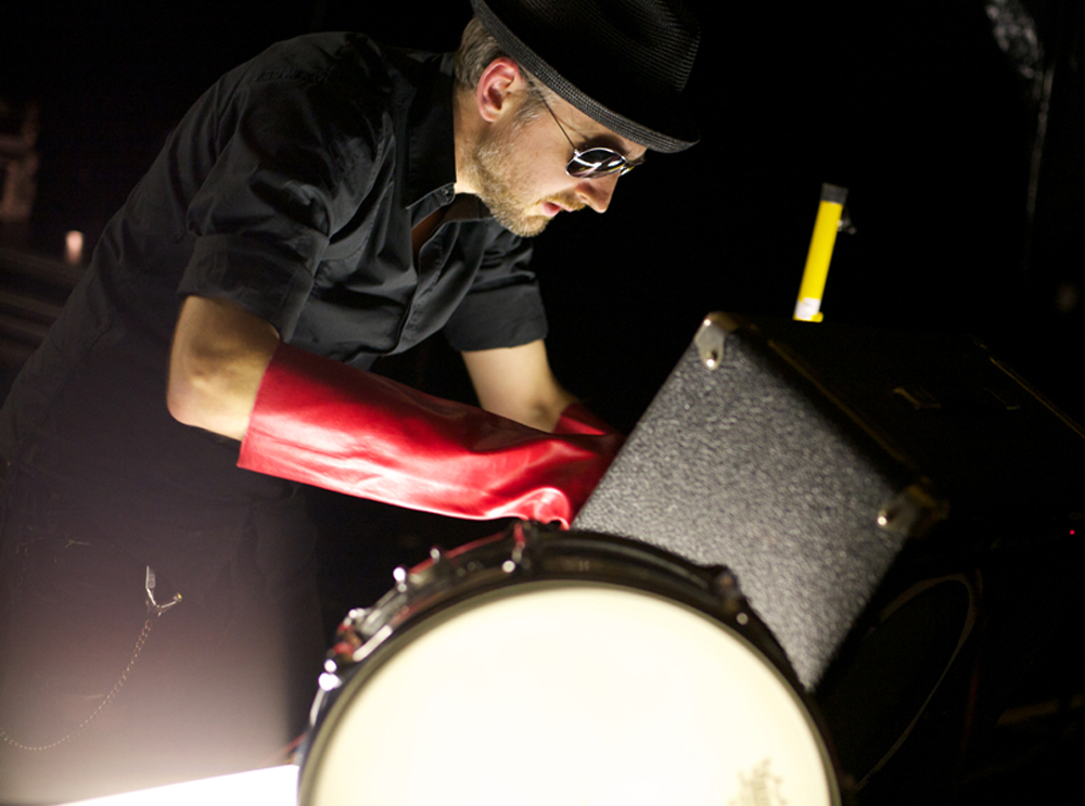 Tim Goldie wearing a red rubber glove, hat and sunglasses looks in an amp case