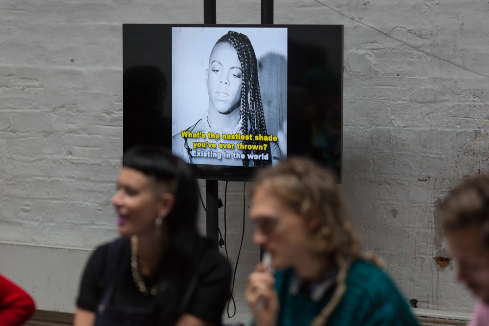 A tv screen with a gif of Juliana Huxtable plays behind people in a workship