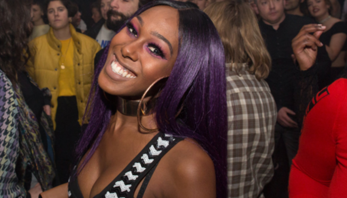 Ahya Simone with purple hair a striped top and big earring smiles hugely