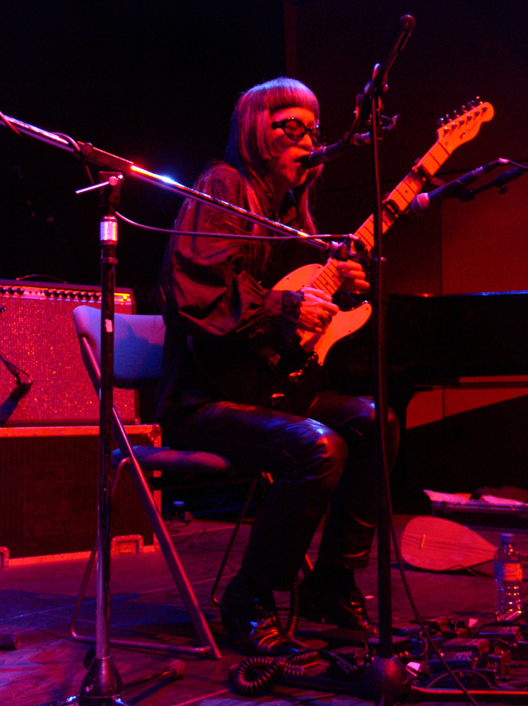 Keiji Haino seated, singing into a microphone while playing a guitar