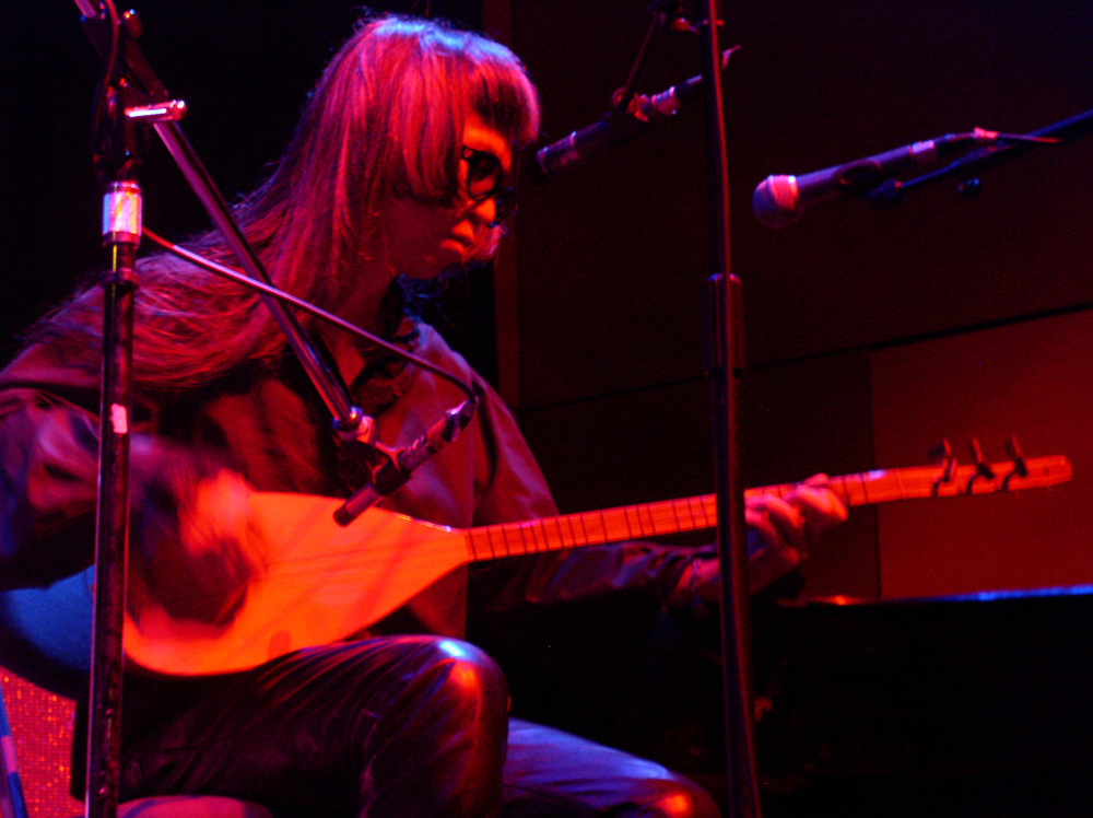 Keiji Haino seated playing a saz, close up in pink light