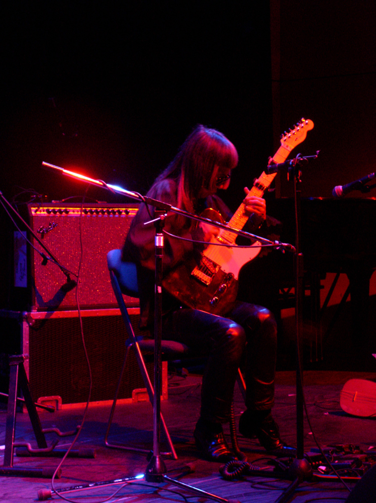 A man seated in pink light playing an electric guitar