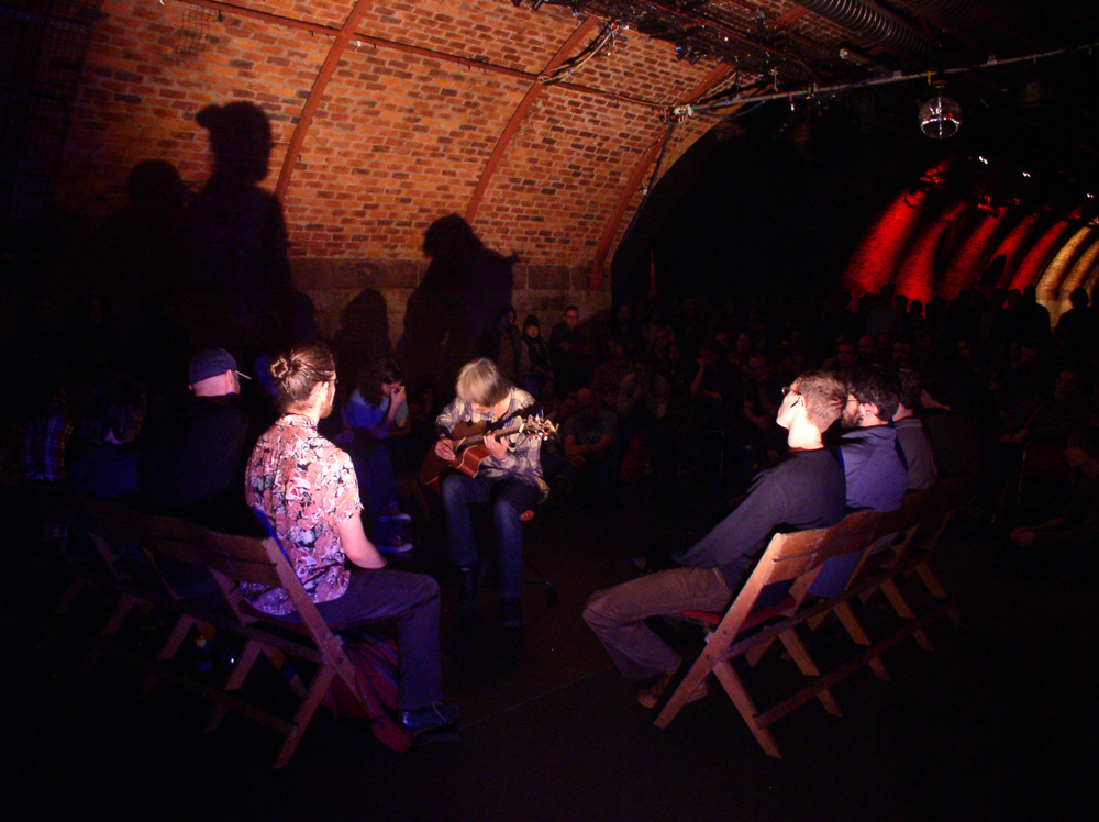 A man playing a guitar framed by starkly lit audience members