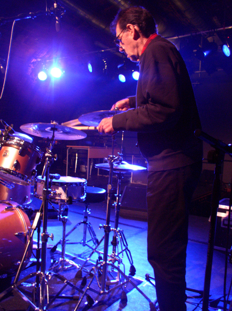 A man approaches a drum kit holding a cymbal