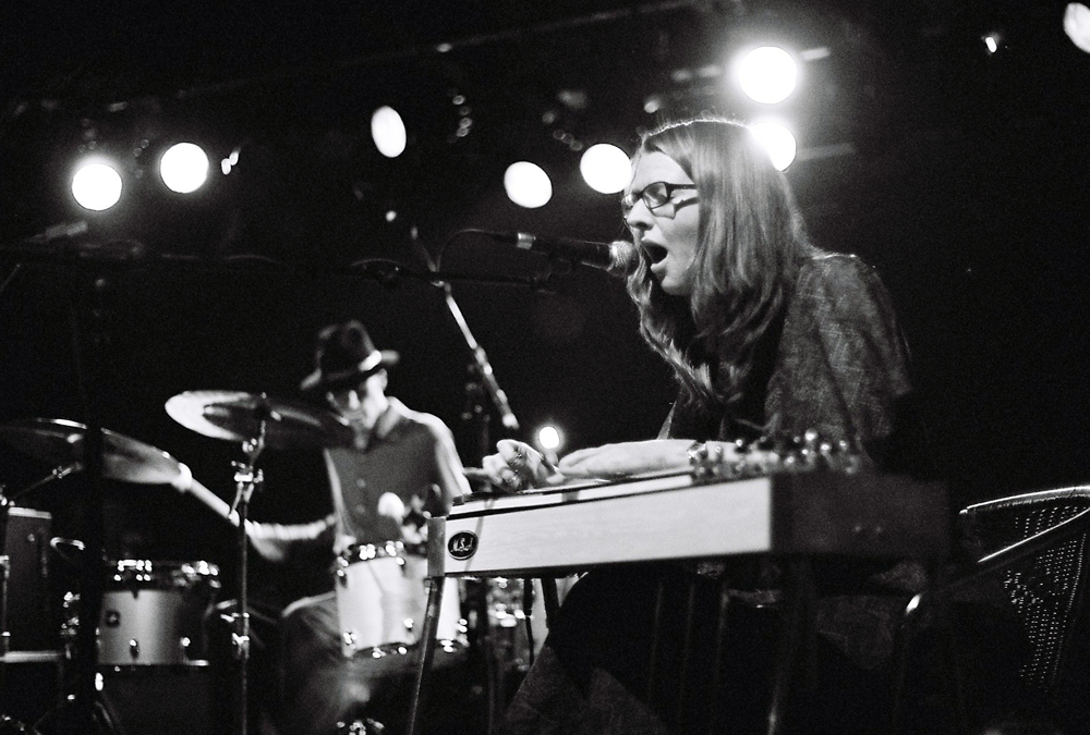 a man playing drums behind a woman playing a pedal steel guitar