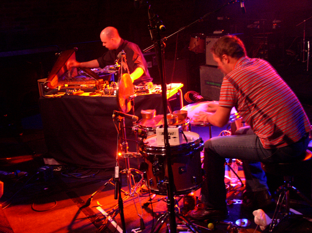 Two men touching carefully a harp and a floor tom in orange light