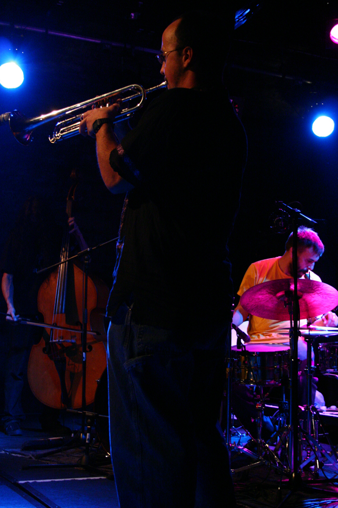 A man plays a trumpet facing away, a drummer and a double bass to the rear