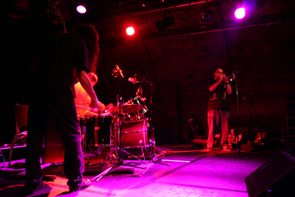 Eye Contact group on stage in pink light, bass, drums, trumpet left to right