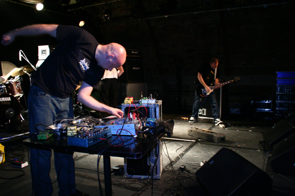 a guitarist and a bald man perform noise music in bright lights
