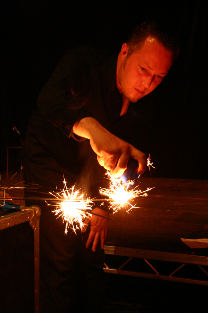 A man's hand reaches forward to light more fireworks, his face is orange 