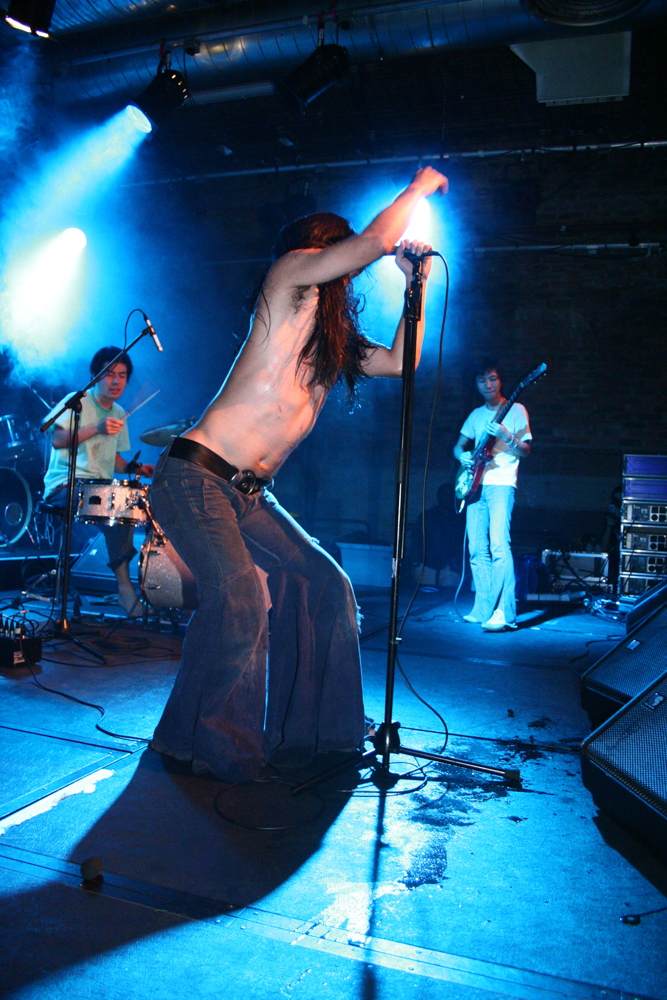 Three men performing rock music, the singer wearing flared trousers