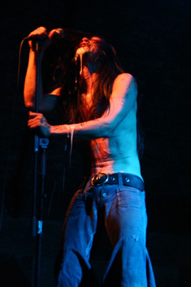 A thin man with scars and vomit on his face holds a microphone
