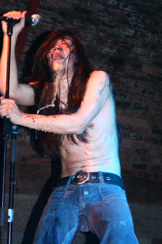A half naked man producing liquids from many orifices holds a microphone 