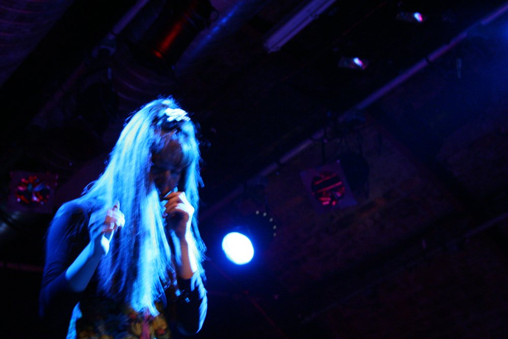 A woman with long hair holds a microphone to her mouth in blue light