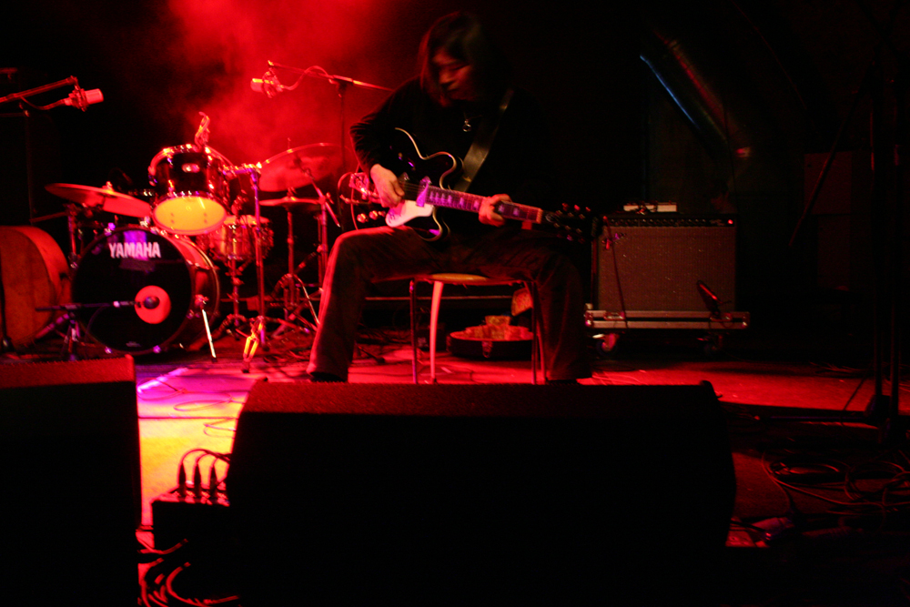A guitarist sitting in front of equipment legs spread with pink cloud of smoke