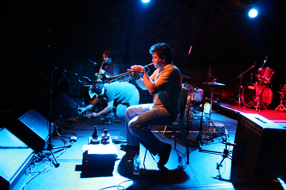 Trio: a man on the floor adjusts a knob, trumpet and sax players on chairs