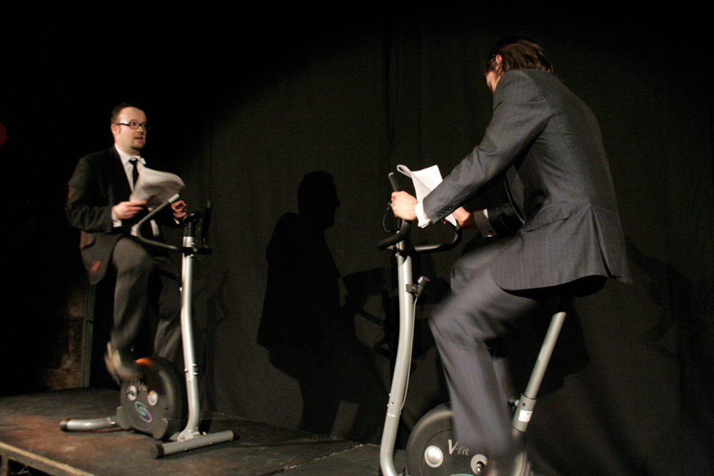 Two men in suits and ties ride exercise bikes whilst reading from books