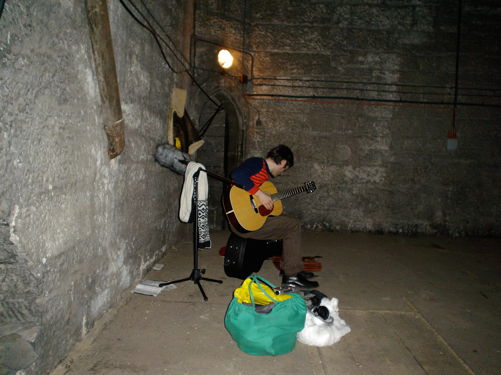 Neil sitting down near a wall of a bell tower holding a guitar