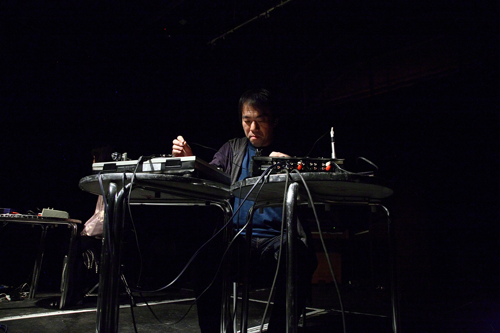Otomo Yoshihide sits at two silver tables playing electronics 