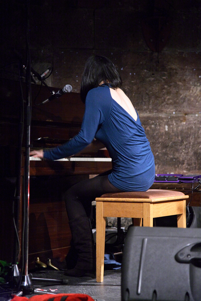 Mico in a blue backless dress sits on a stool as she plays the pianos