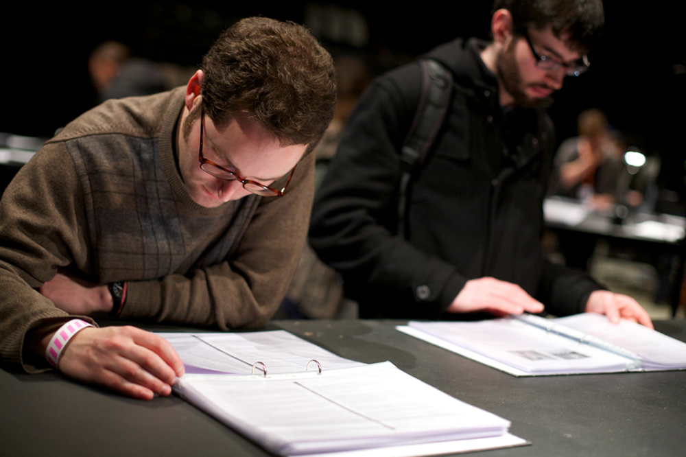 Two people reading documents laid out on a table, both wear glasses