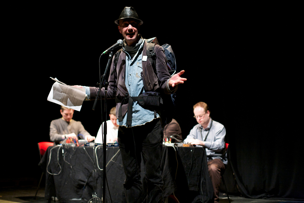 A man with a rucsac and hat reads into a microphone