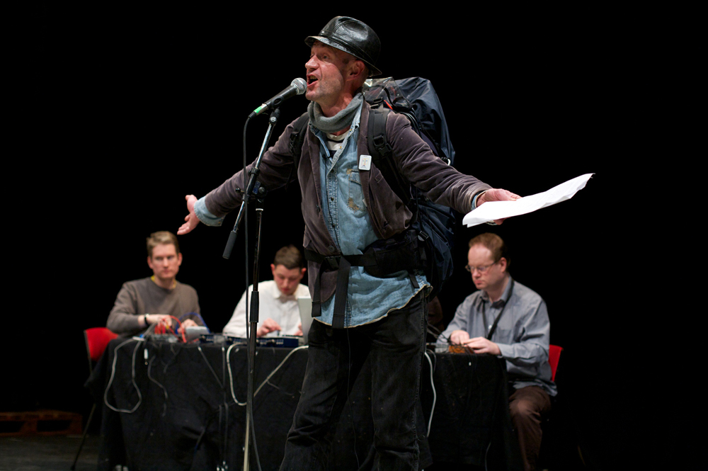 Tam Dean Burn in hat, jacket and rucksack gestures with outstretched arms