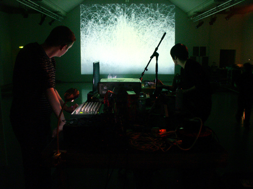 Two performers either side of a projector and desk, a screen in the distance