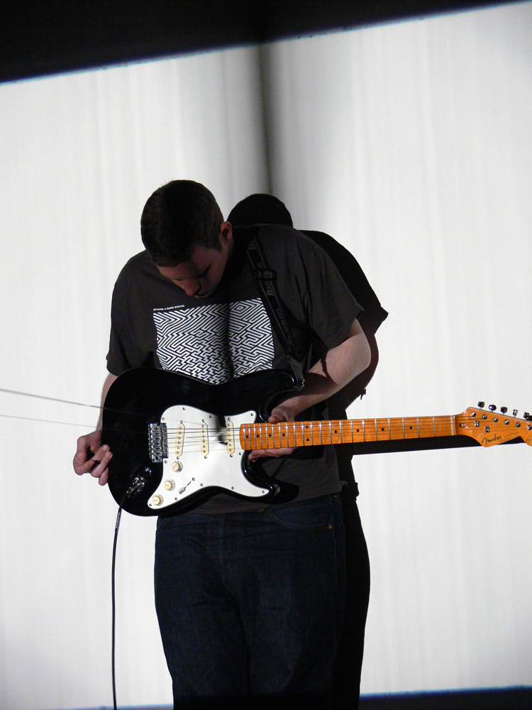 A man standing holding an electric guitar, a band of film looped in the strings