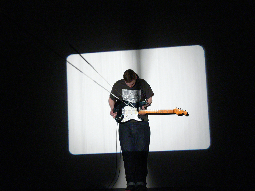 A man standing holding an electric guitar, a band of film looped in the strings