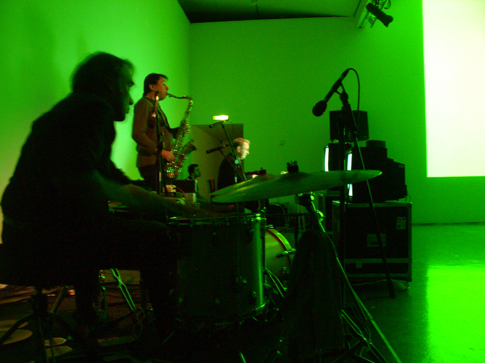 Musicians in green light; drummer, saxophonist, someone in the distance