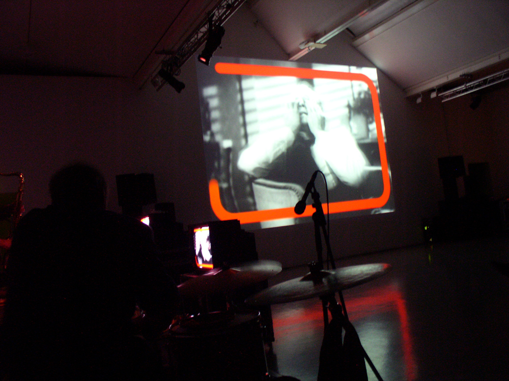 A woman's face and hands framed by orange lines on a screen, musicians nearby